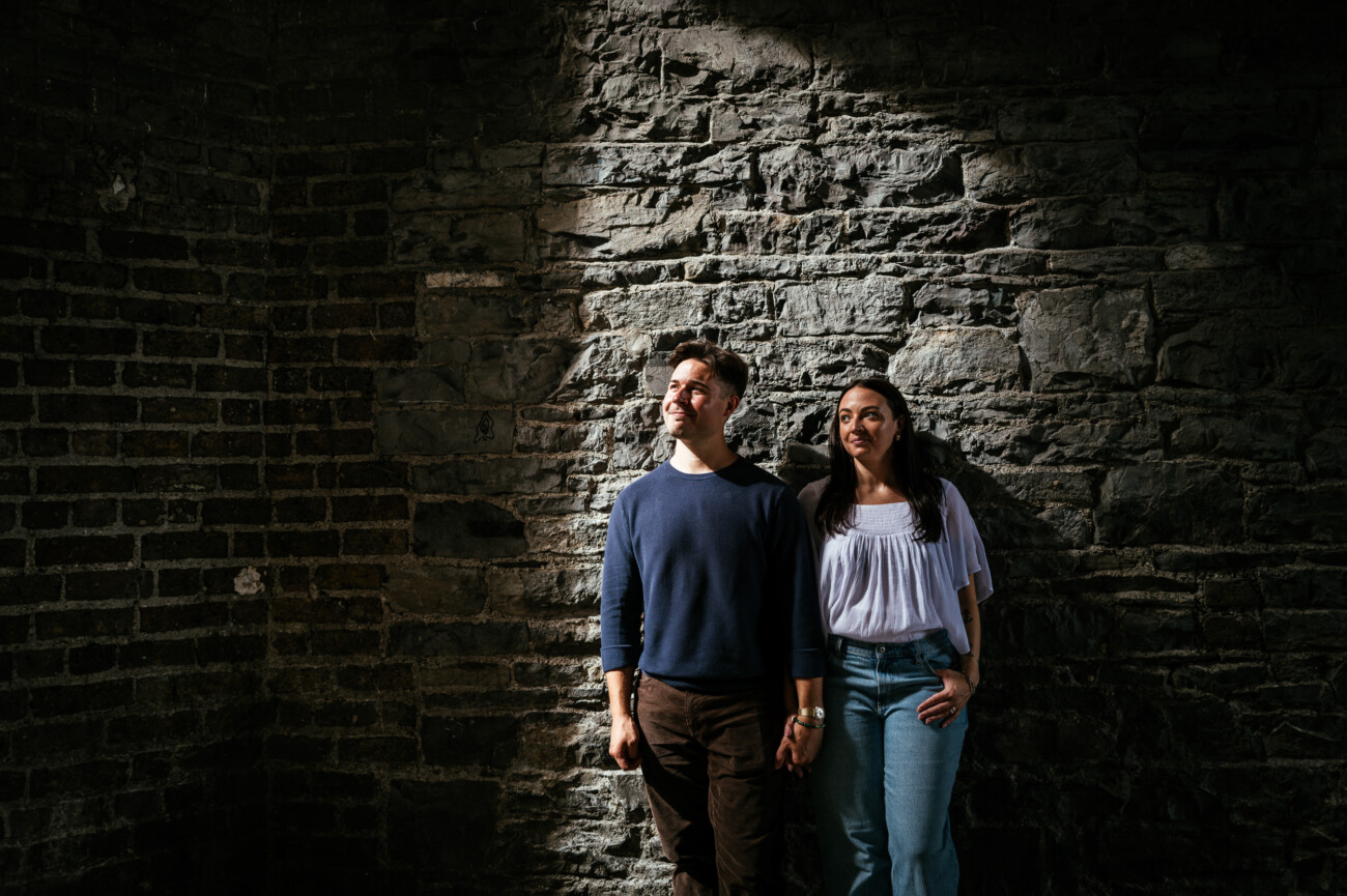 A Secret Proposal at Howth Cliffs & a Private Hour at Howth Castle: Joseph & Shannon's Engagement Shoot 63 A man and a woman stand holding hands against a textured stone wall, illuminated by a shaft of sunlight in an otherwise dimly lit space.