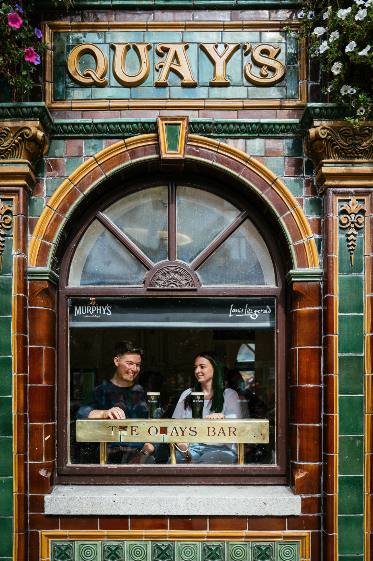 A Secret Proposal at Howth Cliffs & a Private Hour at Howth Castle: Joseph & Shannon's Engagement Shoot 64 Two people sit inside The Quays Bar, seen through a window with tiled green and brown exterior, each with a pint of beer on the table between them.