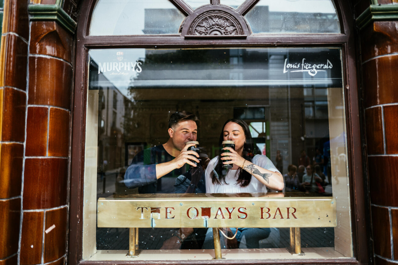 A Secret Proposal at Howth Cliffs & a Private Hour at Howth Castle: Joseph & Shannon's Engagement Shoot 65 A man and woman sit by a window at The Quays Bar, each holding a pint of dark beer and smiling, with the street scene reflected in the glass.