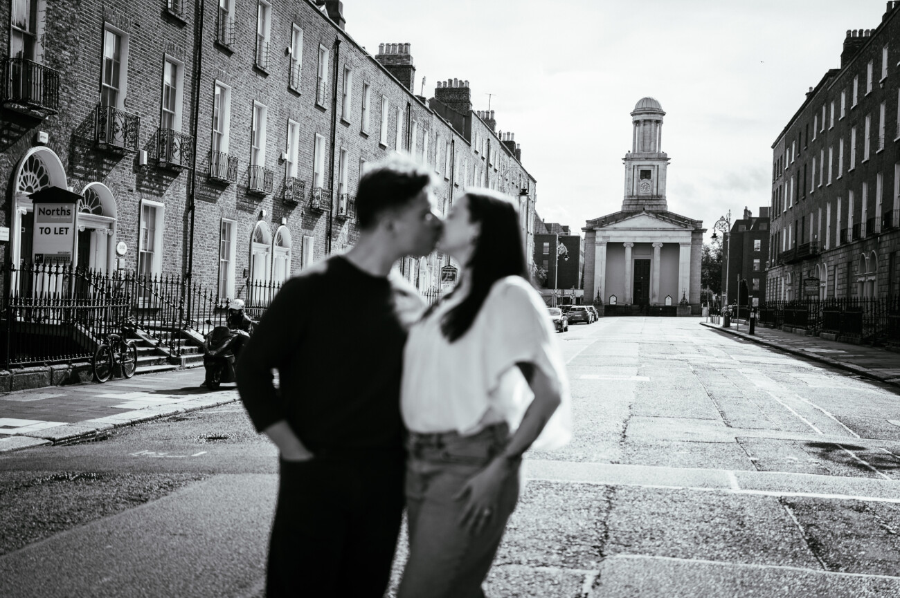 A Secret Proposal at Howth Cliffs & a Private Hour at Howth Castle: Joseph & Shannon's Engagement Shoot 49 A blurred couple stands close together, kissing in the middle of an empty city street lined with brick buildings and a church in the background.