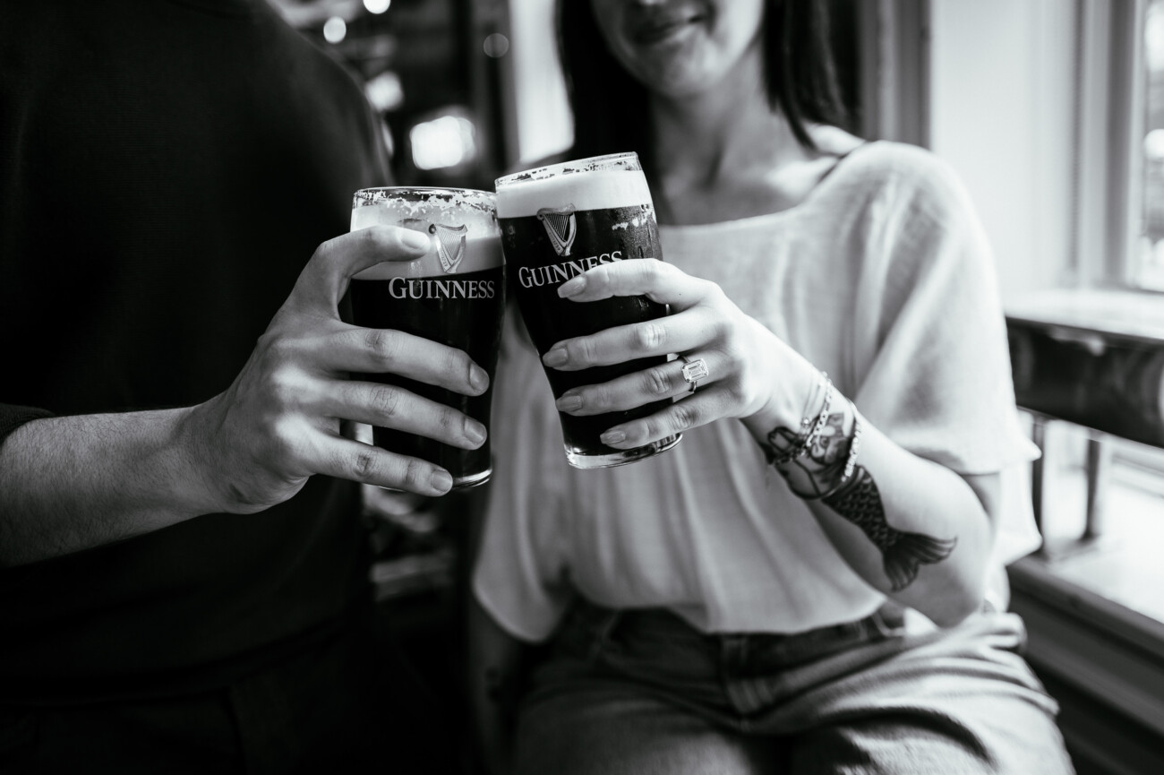 A Secret Proposal at Howth Cliffs & a Private Hour at Howth Castle: Joseph & Shannon's Engagement Shoot 66 Two people clink glasses of Guinness beer together, seated indoors by a window.