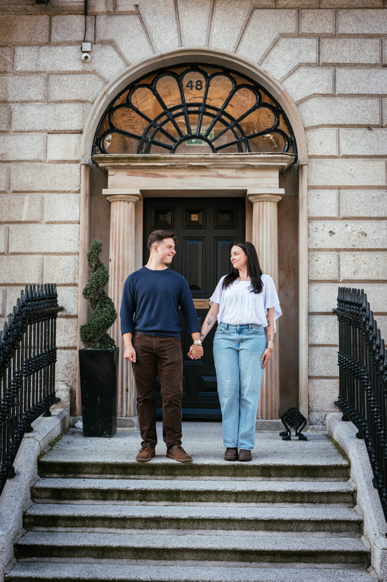 A Secret Proposal at Howth Cliffs & a Private Hour at Howth Castle: Joseph & Shannon's Engagement Shoot 50 A man and woman hold hands while standing on the steps in front of a black door with an arched window above it, outside a stone building.