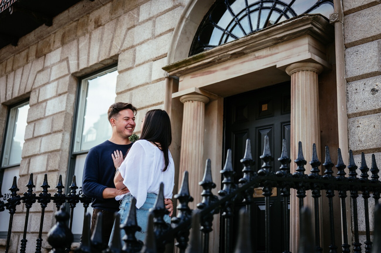 A Secret Proposal at Howth Cliffs & a Private Hour at Howth Castle: Joseph & Shannon's Engagement Shoot 51 A man and woman stand close together and smile outside a stone building with columns, near a black metal fence.