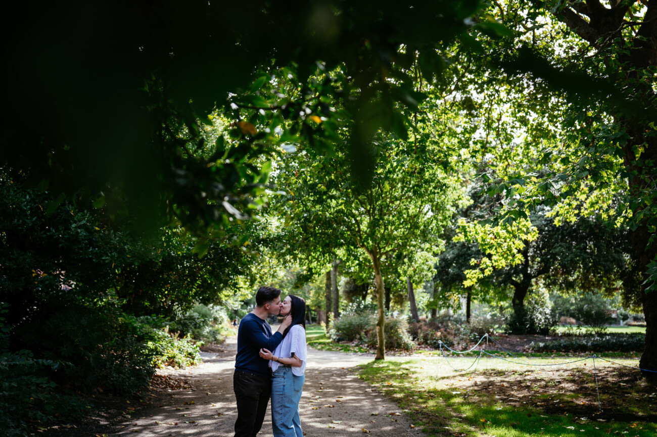 A Secret Proposal at Howth Cliffs & a Private Hour at Howth Castle: Joseph & Shannon's Engagement Shoot 53 A couple snogs while standing on a sunlit path in a park surrounded by lush green trees and foliage.