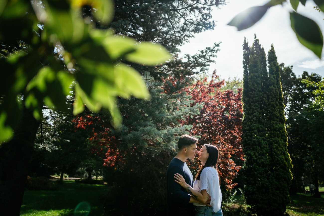 A Secret Proposal at Howth Cliffs & a Private Hour at Howth Castle: Joseph & Shannon's Engagement Shoot 54 A couple stands close together in a park, surrounded by trees and greenery, with sunlight filtering through the leaves.