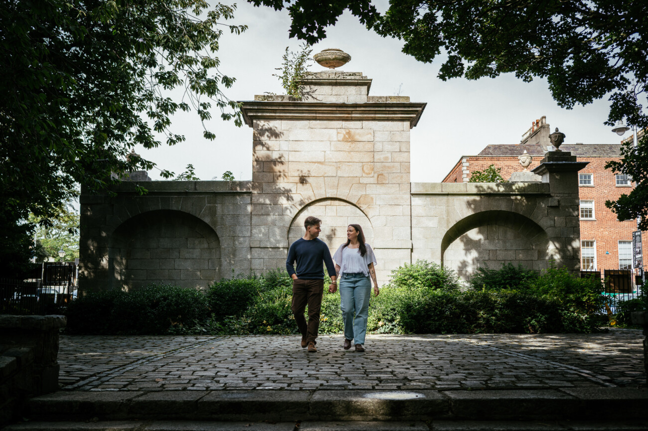 A Secret Proposal at Howth Cliffs & a Private Hour at Howth Castle: Joseph & Shannon's Engagement Shoot 55 A man and woman walk hand in hand on a cobbled path in front of a large stone structure with greenery and trees around them.
