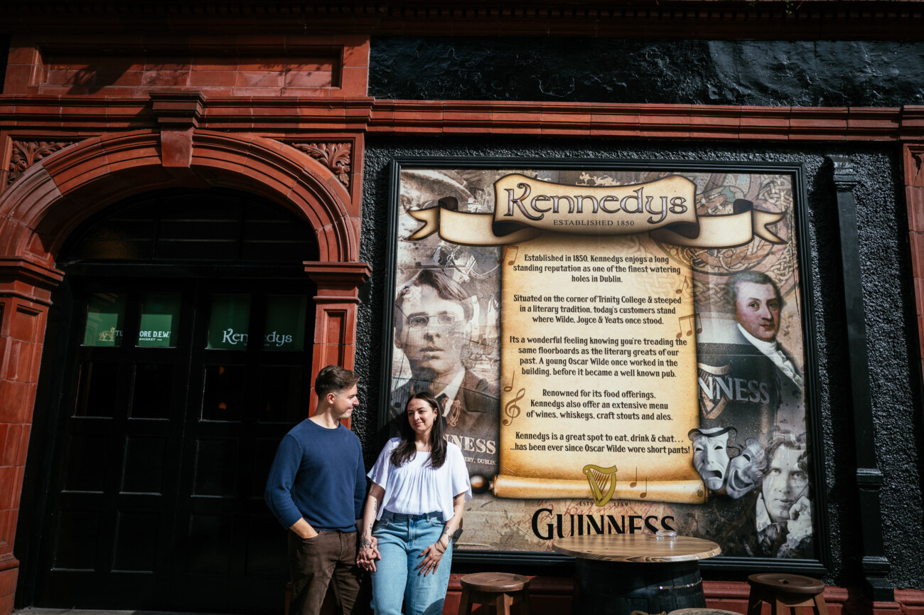 A Secret Proposal at Howth Cliffs & a Private Hour at Howth Castle: Joseph & Shannon's Engagement Shoot 56 A couple stands holding hands in front of Kennedy’s pub, with a large Guinness-themed mural and historic portraits displayed on the wall beside them.